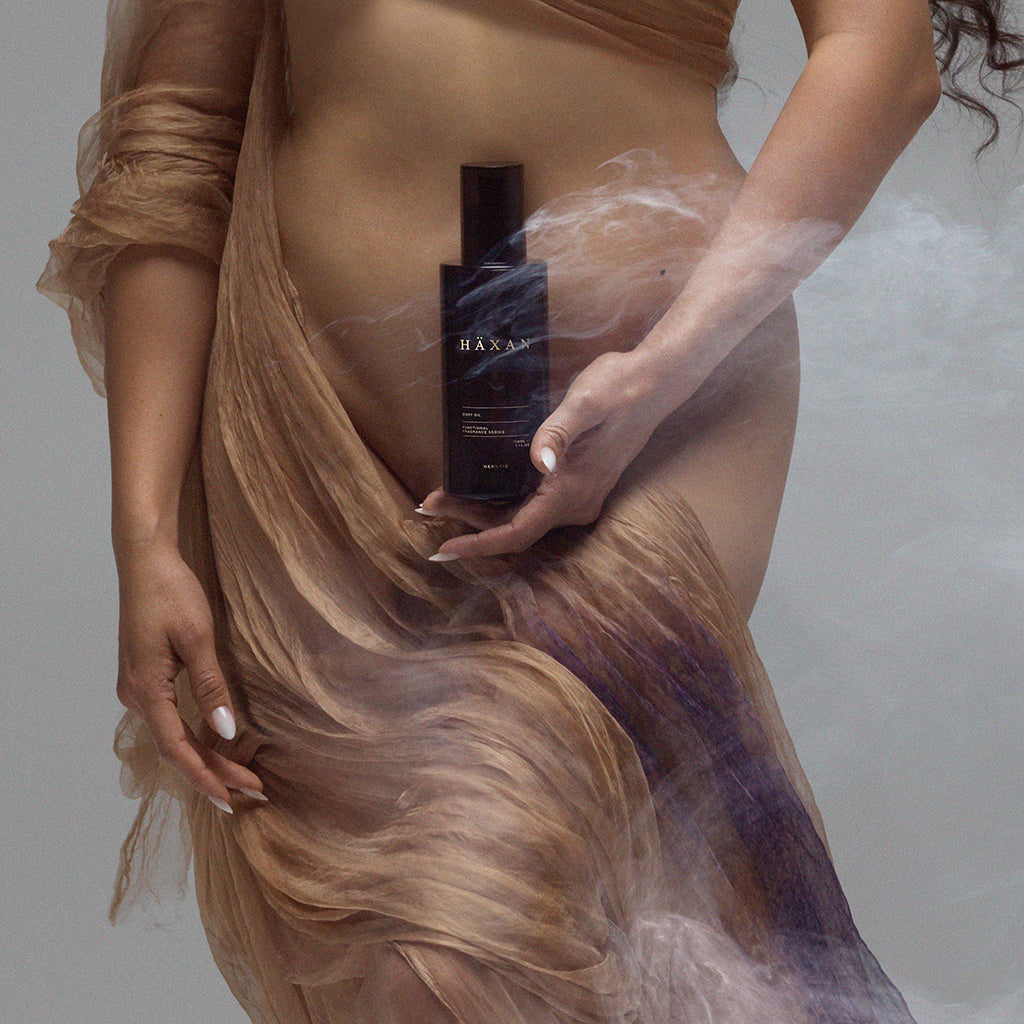 A woman in flowing sheer beige fabric holds a dark rectangular bottle labeled Haxan against a smoky gray studio background.