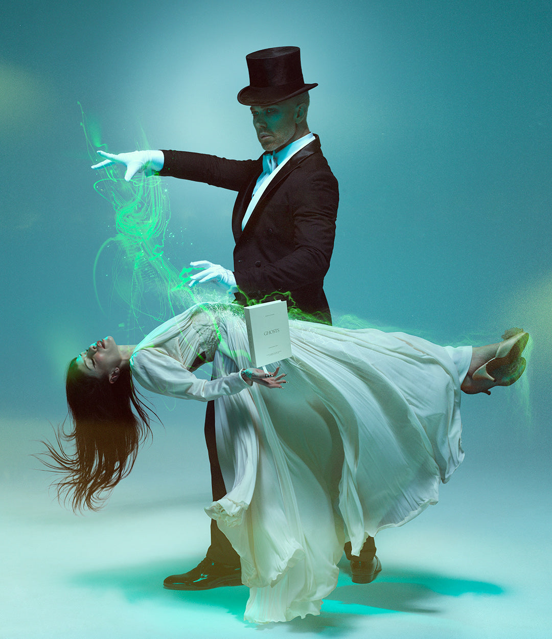 A man in a black tuxedo and top hat stands behind a woman in a flowing white gown who is leaning back dramatically, with glowing green energy effects and a white book in the scene against a blue studio background.