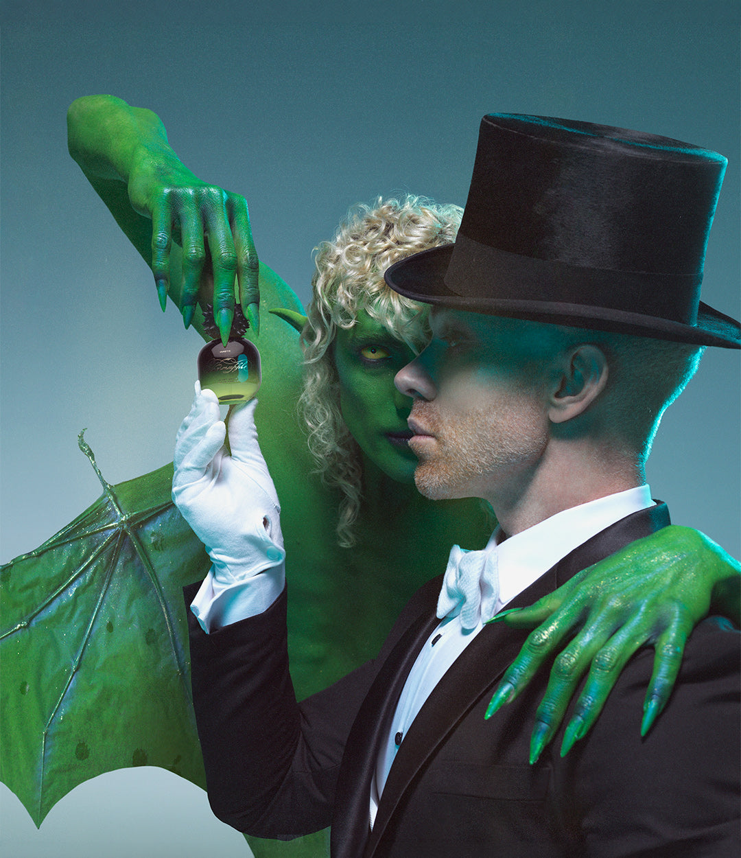 A theatrical photograph featuring a man in a black tuxedo and a creature with green skin and long green fingernails, holding a small glass bottle.